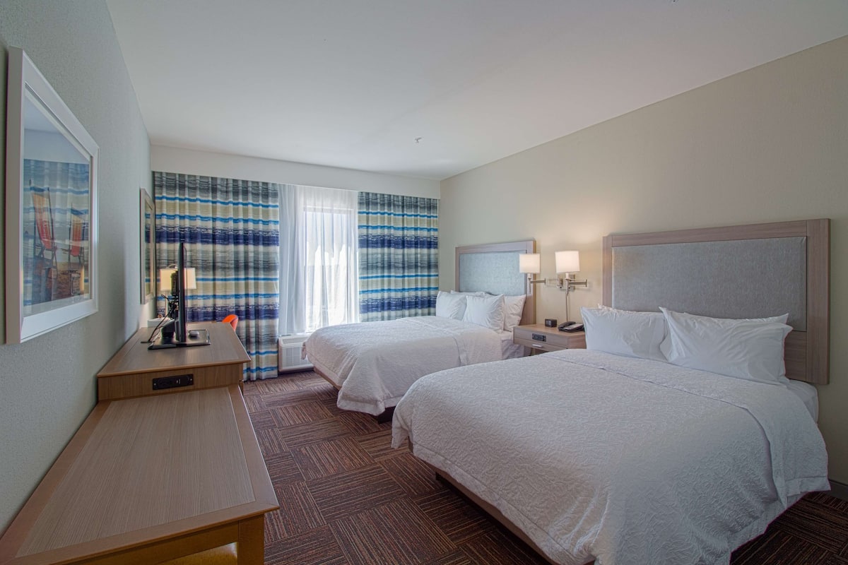 A hotel room with two beds, a desk, a chair, a lamp, and a window with striped curtains.