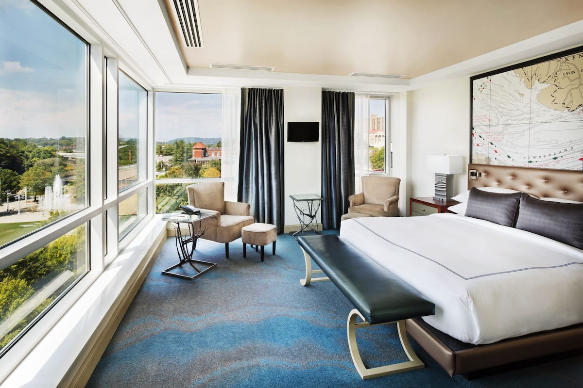 A hotel room with a large bed, a bench, two armchairs, a small table, and a TV.