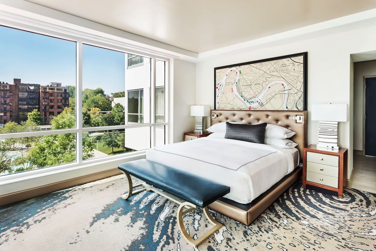 A hotel room with a large window, a bed with a headboard, a bench, a nightstand, and a map on the wall.