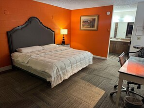 Habitación de hotel con cama, mesita de noche con lámpara, una obra de arte en la pared y vistas al baño y la cocina a través de una puerta abierta.