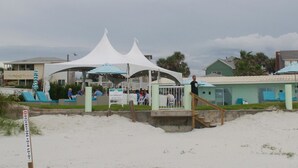 Un complexe hôtelier en bord de mer, avec des tentes blanches, des chaises longues bleues et un panneau indiquant qu’il est interdit de se baigner au-delà d’un certain point.