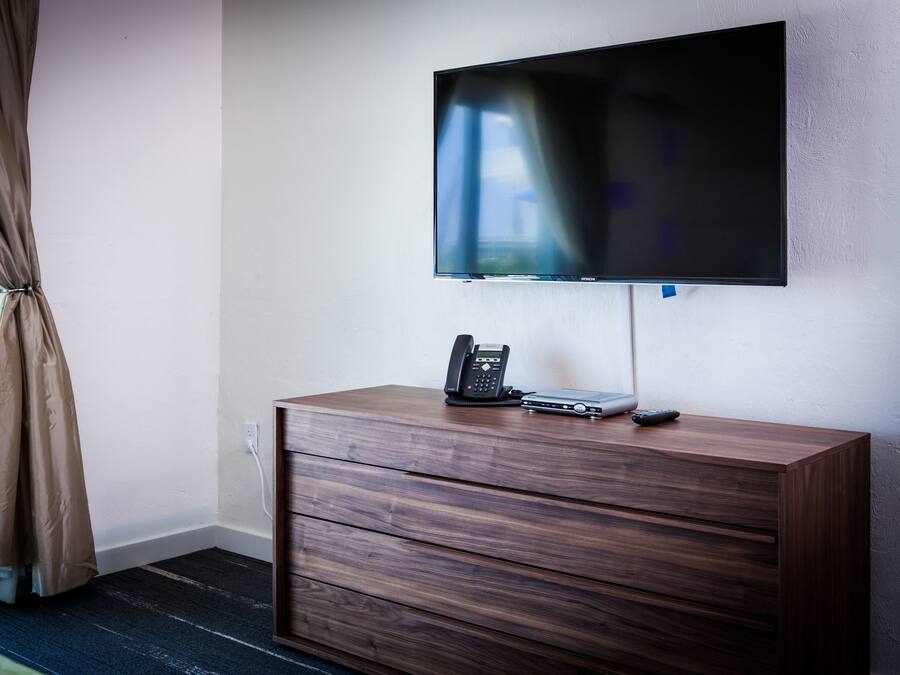 Ein Hotelzimmer mit einem an der Wand befestigten Flachbildfernseher, einer Holzkommode mit Telefon und Fernbedienung sowie einem Fenster mit Vorhängen.