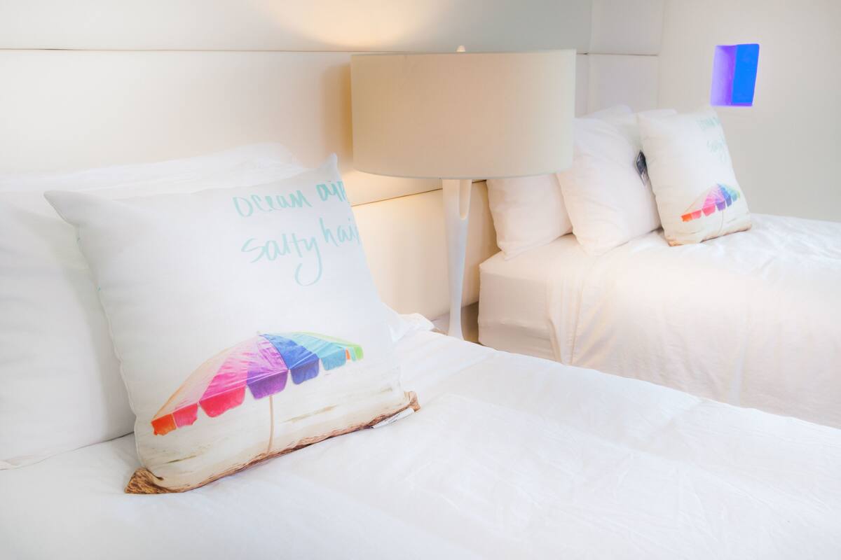 A hotel room with two beds, each with white bedding and pillows. One pillow has text and a colorful design.
