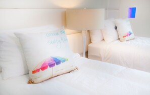 Quarto de hotel com duas camas, cada uma com roupa de cama e almofadas brancas. Uma almofada tem texto e um design colorido.