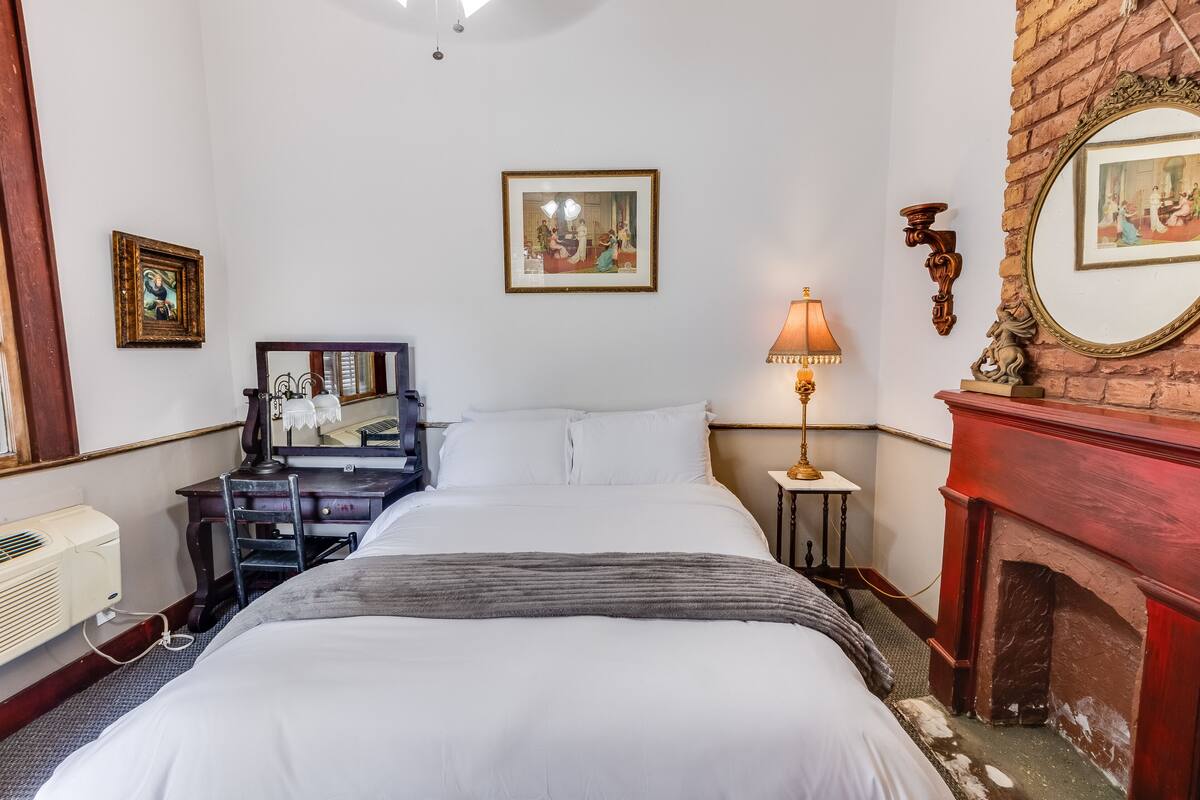 Quarto de hotel com cama, escrivaninha, cadeira, lareira e quadros nas paredes.