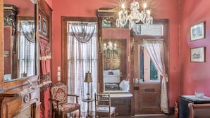 A room with a wooden floor, a chandelier, a mirror, a chair, and a bed with a red curtain. - Royal Barracks Guest House (New Orleans)
