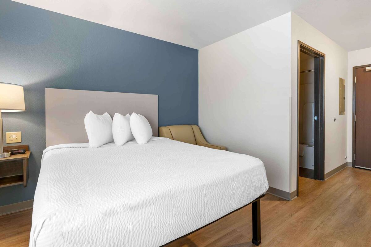 Una habitación de hotel con una cama grande, una mesita de noche, una lámpara y una puerta que da al baño.