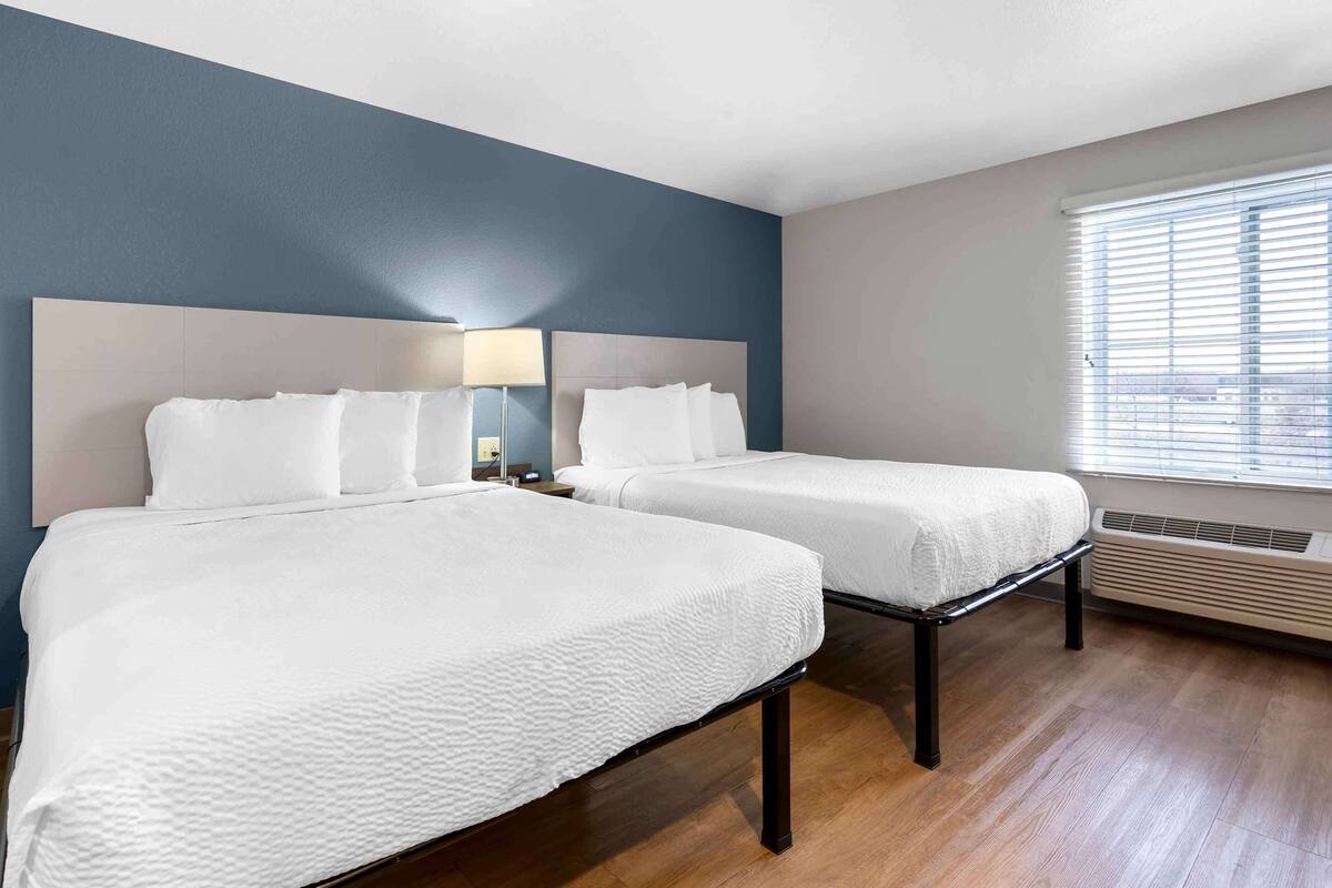 Una habitación de hotel con dos camas, un ventanal con persianas, un aire acondicionado montado en la pared y una mesita de noche con lámpara.
