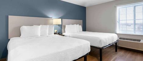 A hotel room with two beds, a large window with blinds, a wall-mounted air conditioning unit, and a nightstand with a lamp.