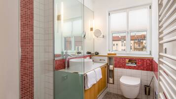 A bathroom with a glass shower enclosure, a white sink, a toilet, and a window with a view of buildings.