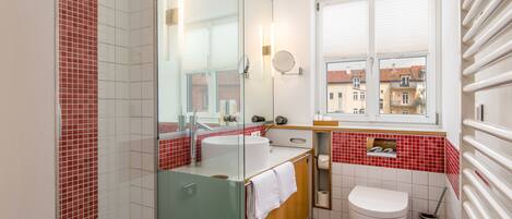 A bathroom with a glass shower enclosure, a white sink, a toilet, and a window with a view of buildings.