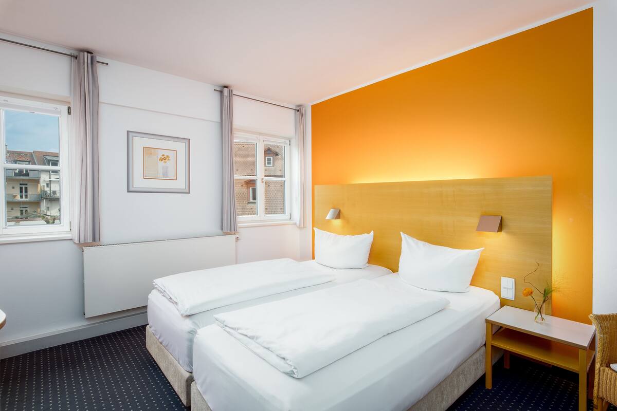 A hotel room with two beds, a window with curtains, a framed picture on the wall, and a nightstand with a vase.