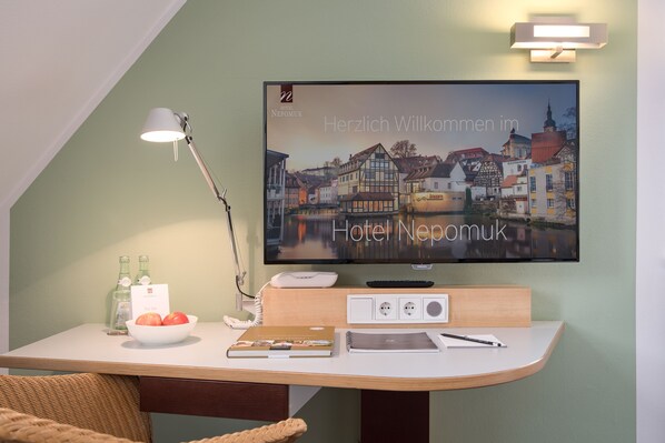 A hotel room with a flat-screen TV displaying a welcome message, a desk with a lamp, a bowl of fruit, and a notebook. - Hotel Nepomuk (Bamberg)