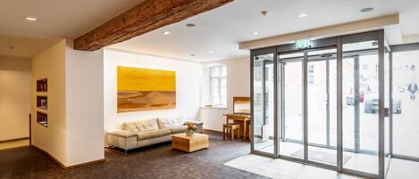 A modern lobby with a wooden ceiling beam, a large glass door entrance, a sofa, a coffee table, and a painting on the wall.