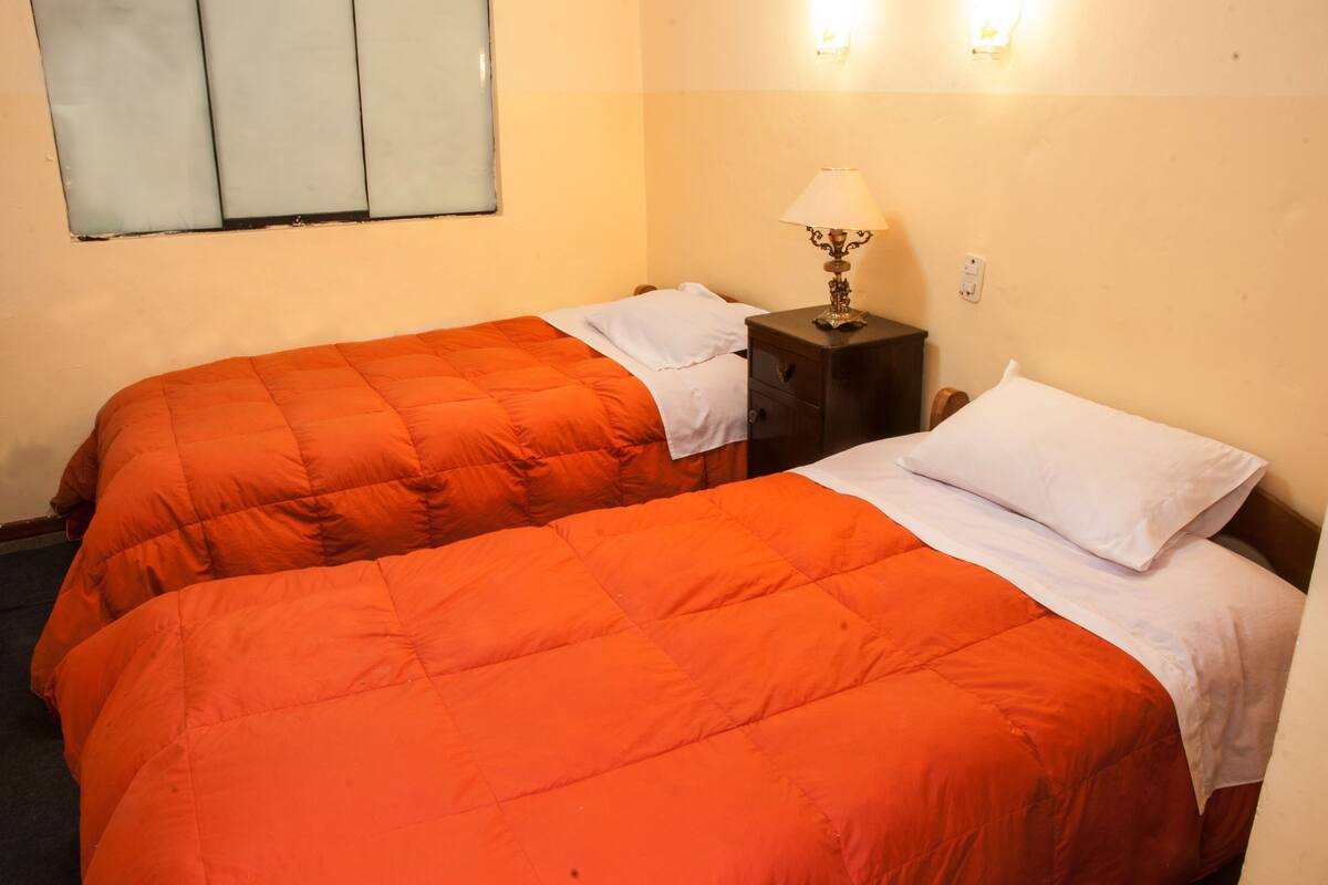 A room with two beds, each with an orange quilt, a nightstand with a lamp, and a wall-mounted light fixture.
