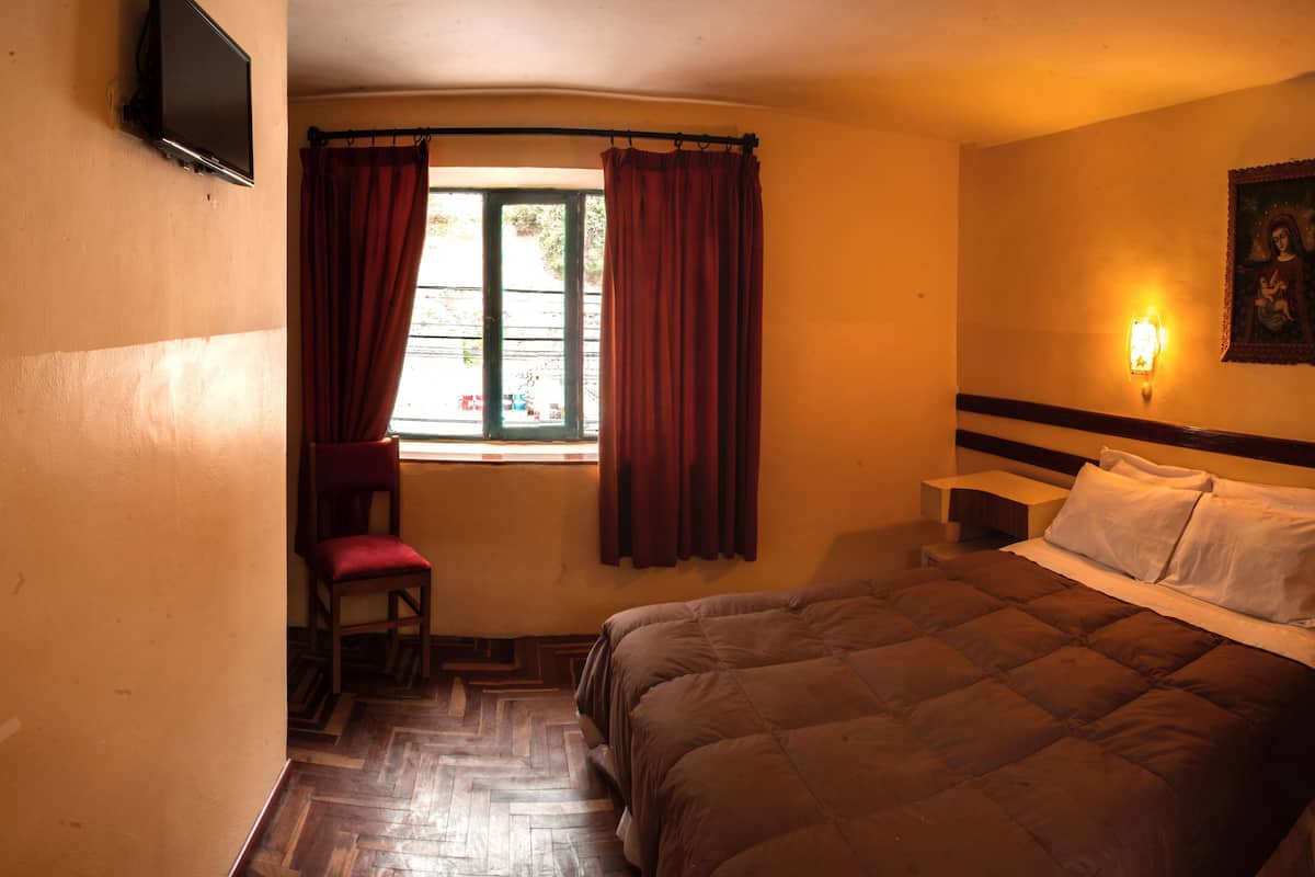 A bedroom with a bed, a nightstand, a chair, a window with curtains, and a wall-mounted light.