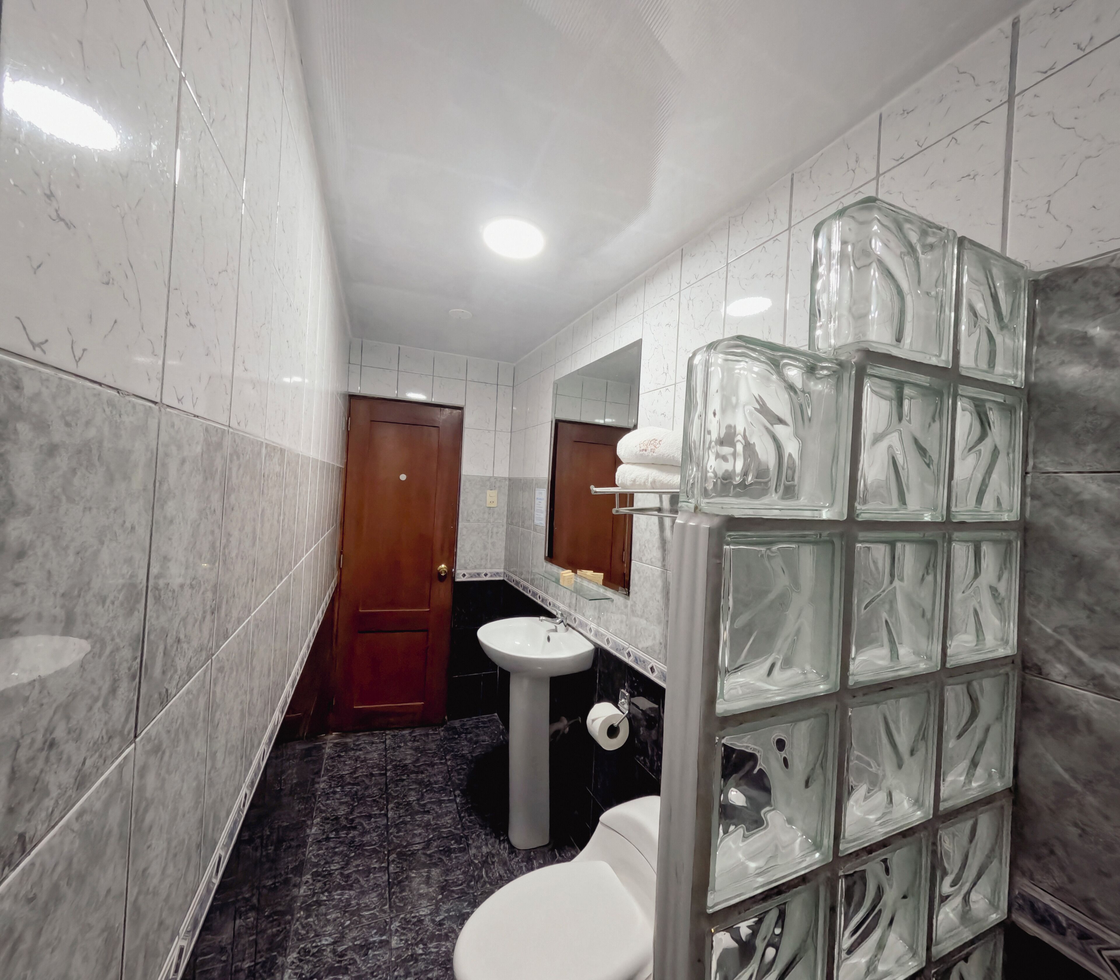 A bathroom with marble tiles, a wooden door, a white sink, a toilet, and a glass shower enclosure.