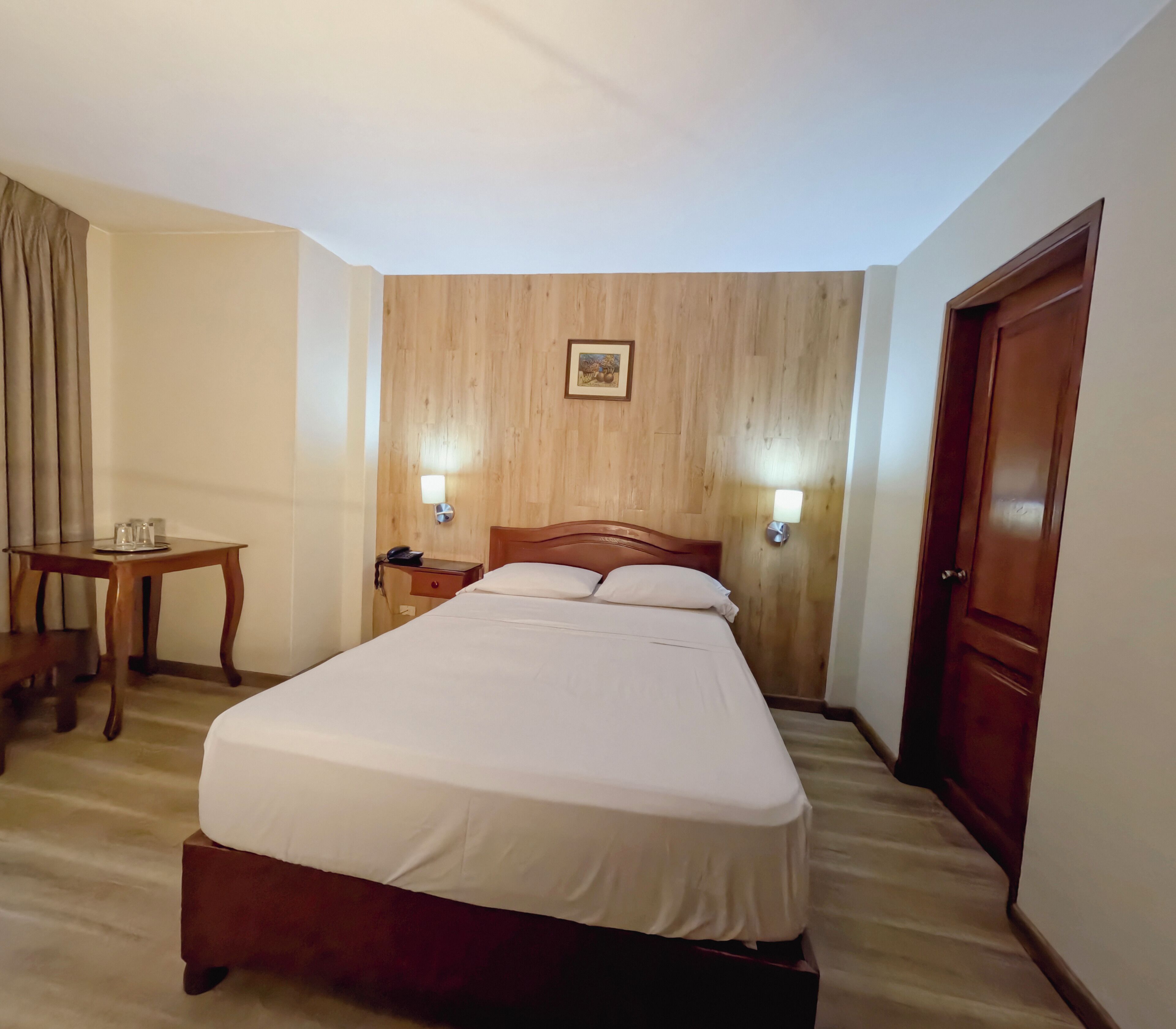 A hotel room with a bed, bedside table, lamp, and a wooden wall panel.