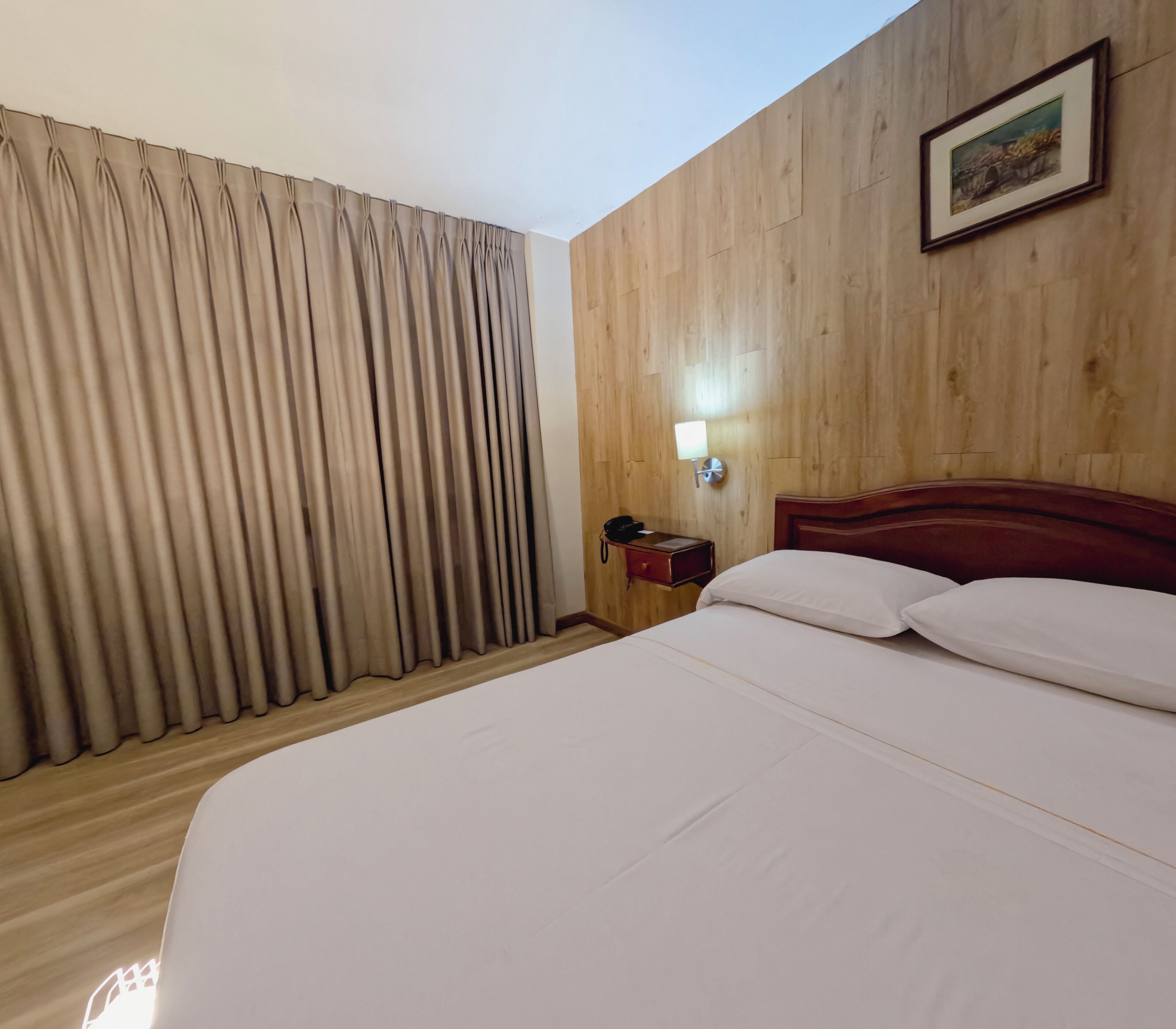 Habitación de hotel con pared de madera, cama con ropa blanca, mesita de noche con lámpara y ventana con cortinas.