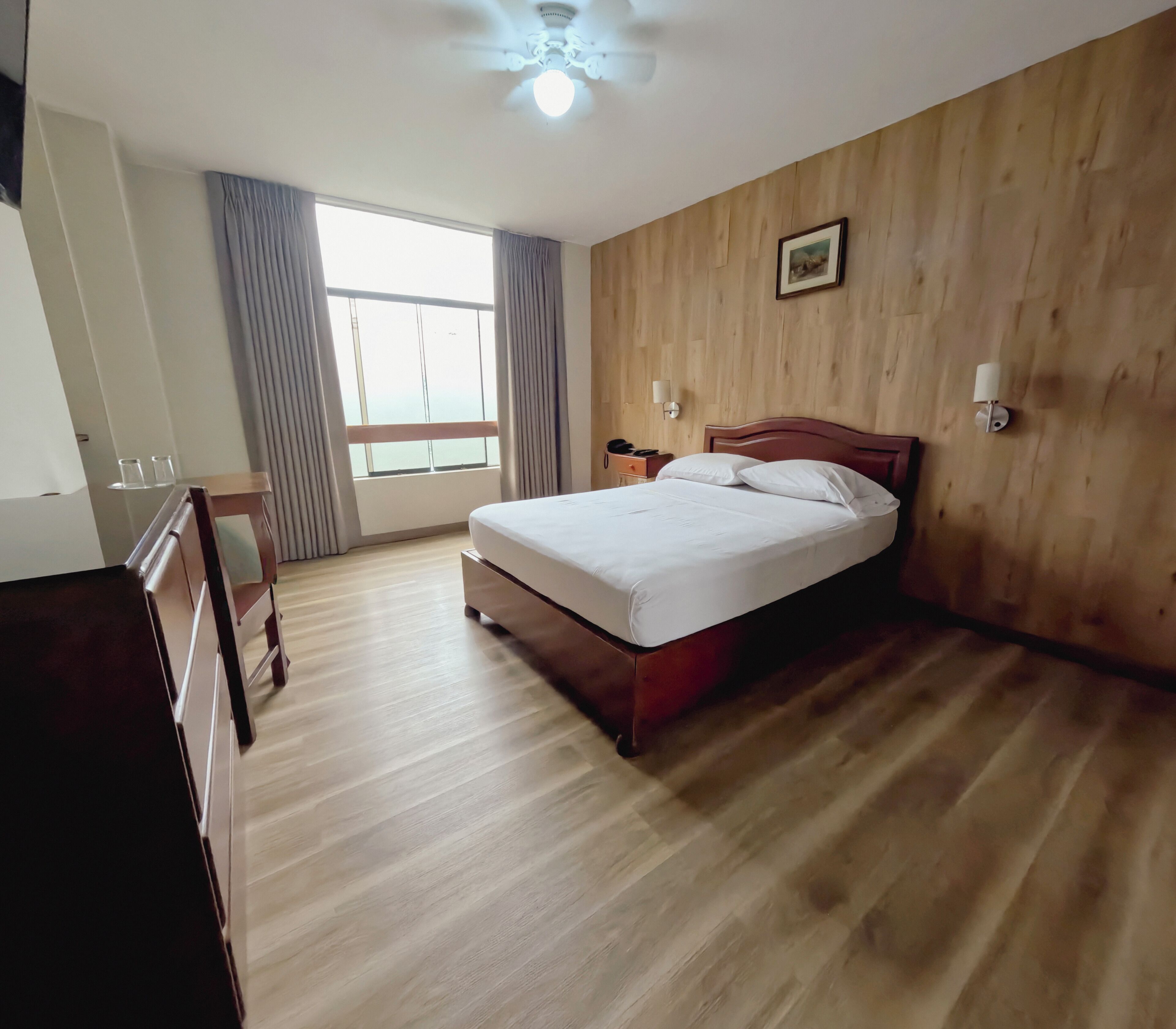 A hotel room with a wooden floor, a bed with white bedding, a nightstand with a phone, a ceiling fan, and a window with curtains.