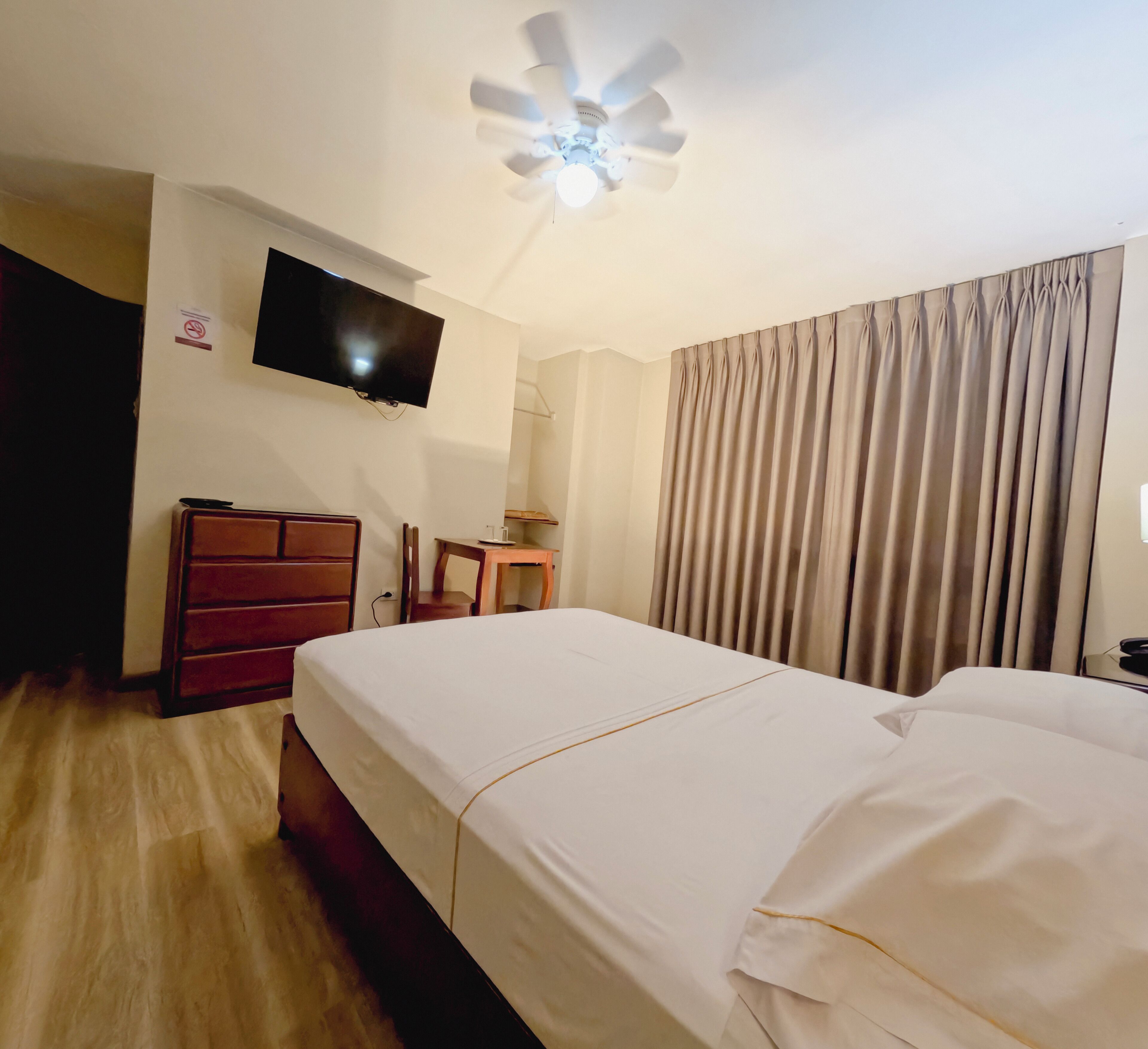 Habitación de hotel con cama, televisión montada en la pared, escritorio, silla y ventilador de techo.