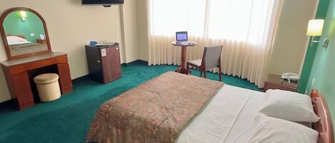 Een hotelkamer met een bed, een bureau met een laptop, een televisietoestel, een plafondventilator en een raam met gordijnen.