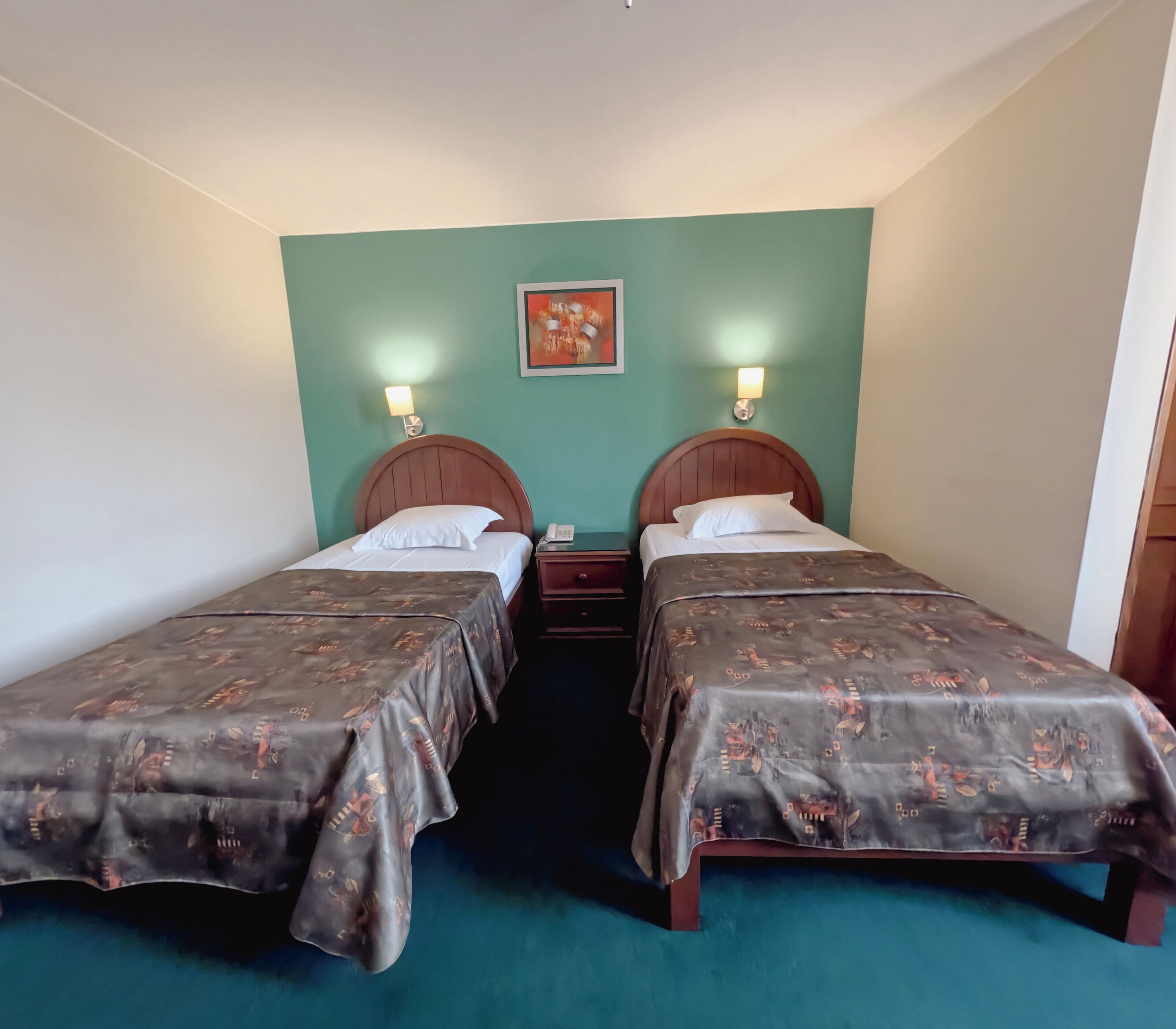 A hotel room with two single beds, each with a wooden headboard and a patterned bedspread. There are bedside tables with lamps and a small painting on the wall above the beds.