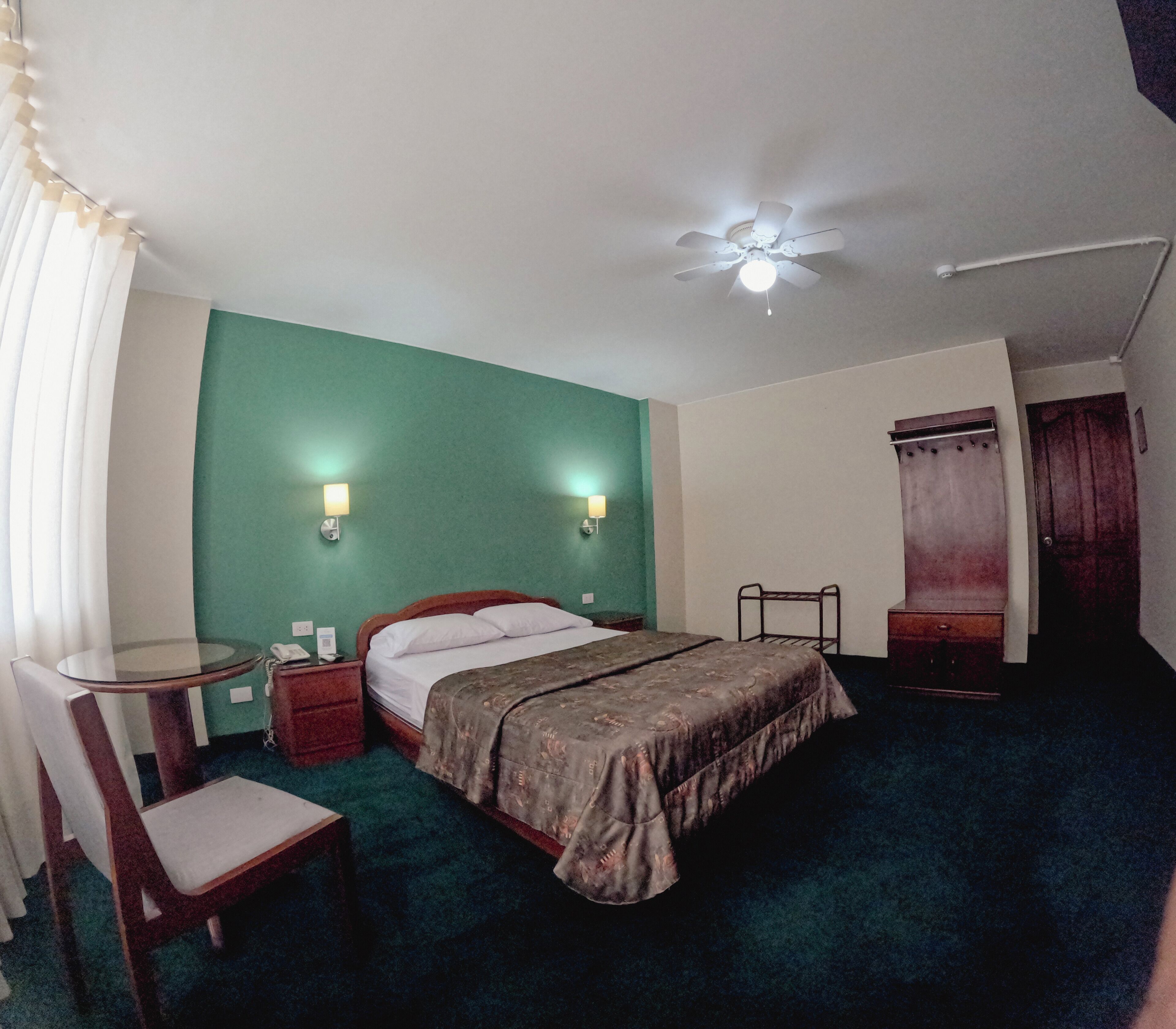 A hotel room with a bed, bedside tables, a chair, a desk, a ceiling fan, and a window with curtains.
