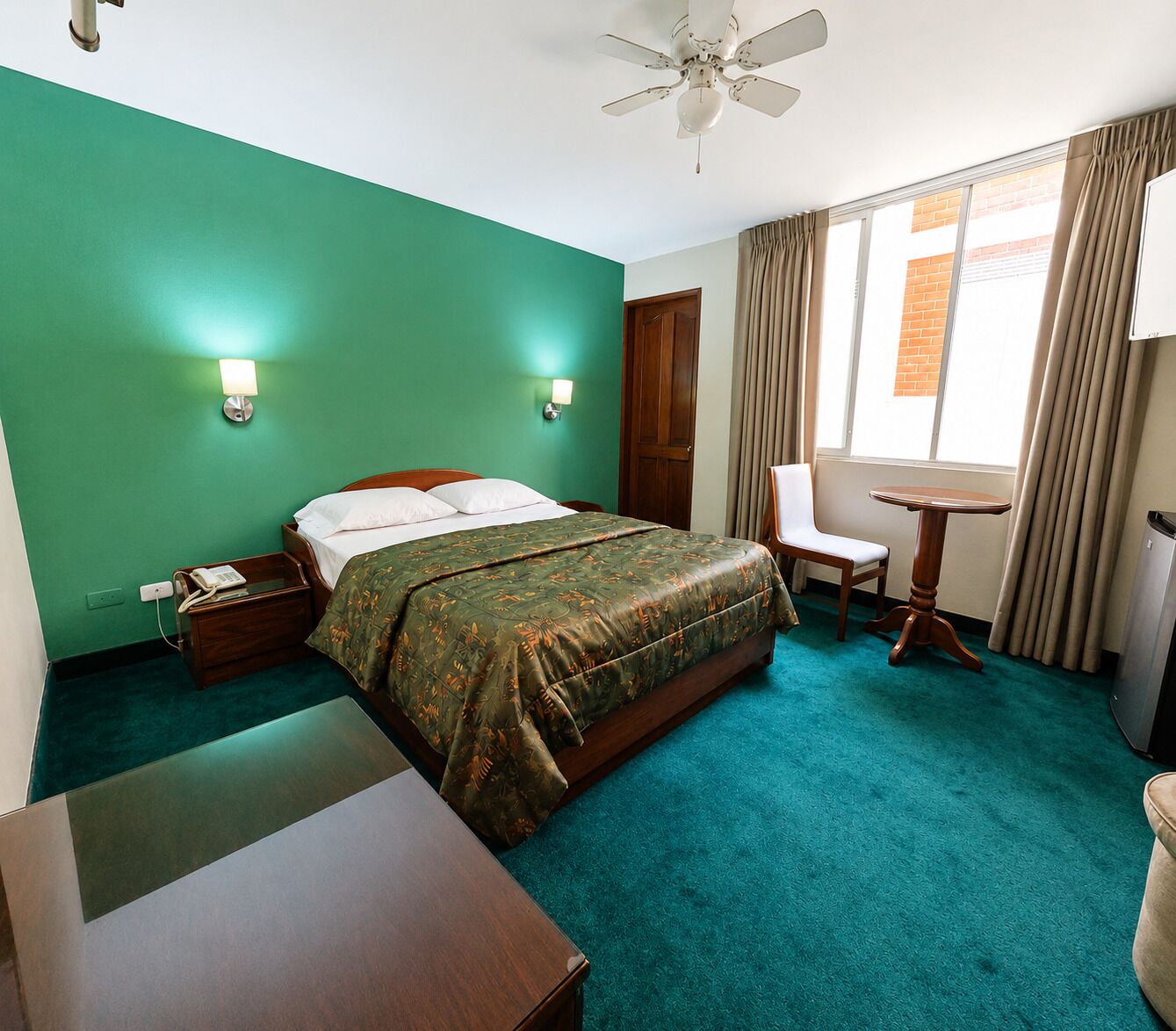 Une chambre d’hôtel équipée d’un lit, d’un bureau, d’une chaise, d’une table, d’un ventilateur de plafond et d’une fenêtre avec des rideaux.