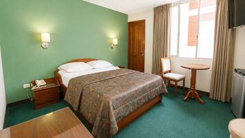 A hotel room with a bed, bedside table, chair, small table, and a television.