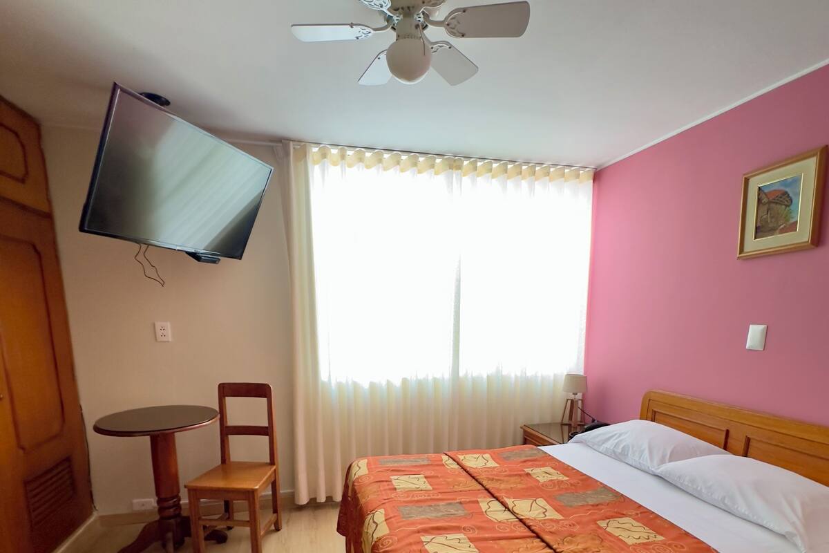Ein Hotelzimmer mit einem Bett, einem Schreibtisch, einem Sessel, einem an der Wand montierten Fernseher und einem Deckenventilator.