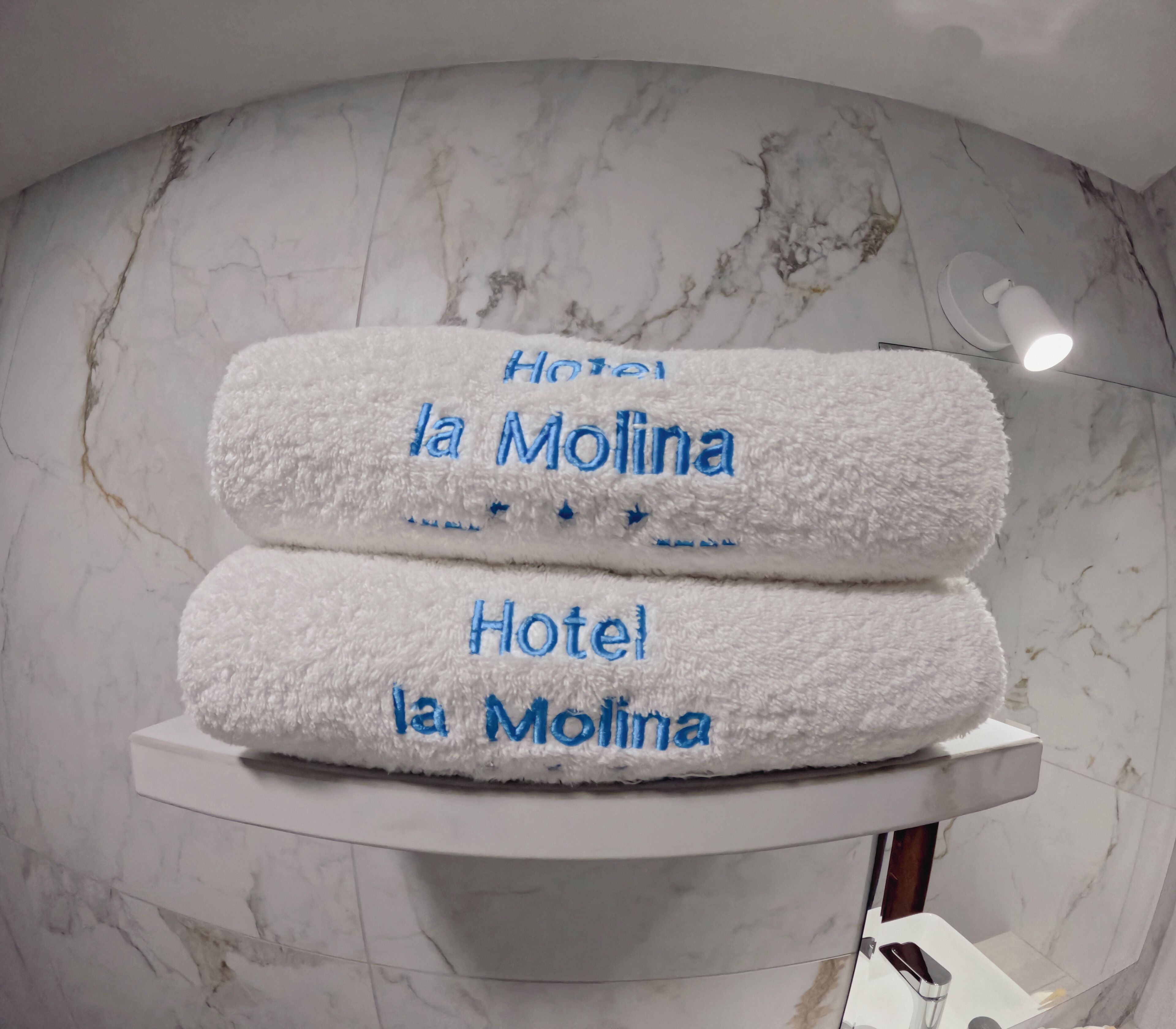 Two white towels with 'Hotel la Molina' embroidered in blue, placed on a white pedestal in a bathroom with marble walls.