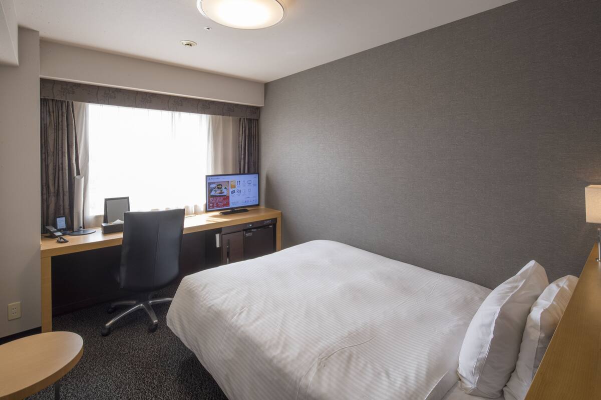 A hotel room with a bed, desk, chair, and a computer monitor.
