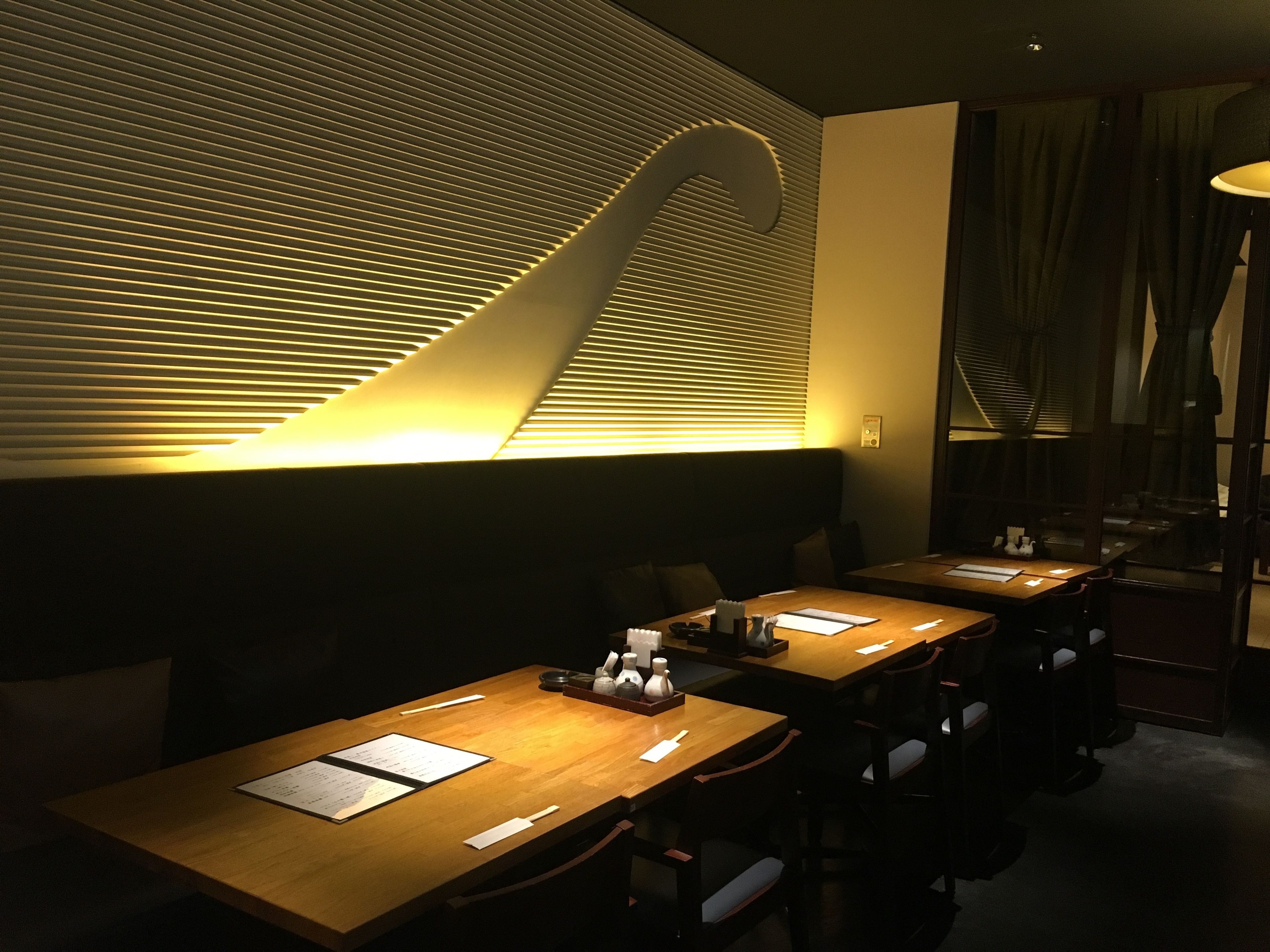 Um restaurante com interior moderno, com mesas de madeira compridas e um design de teto único com linhas inclinadas.