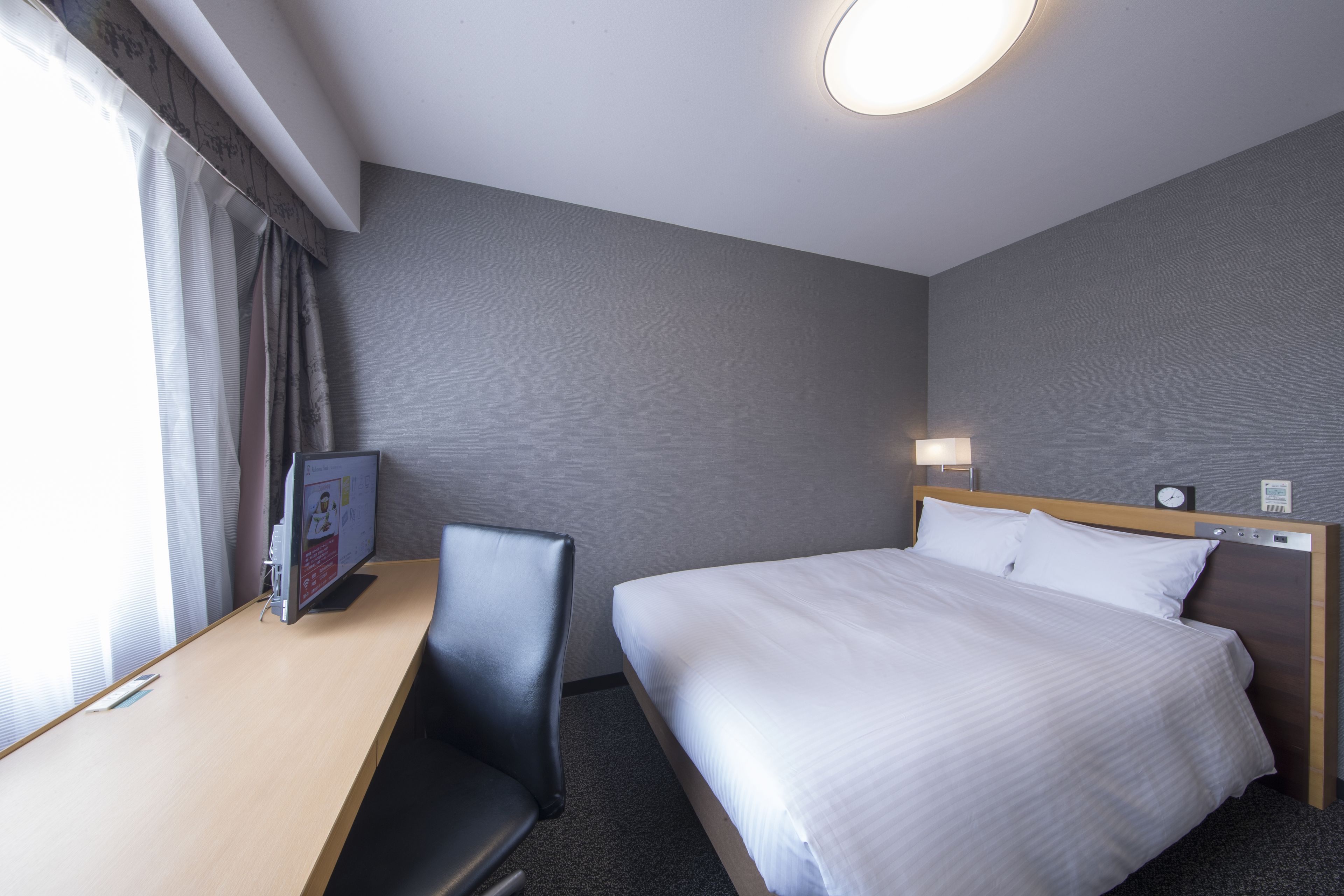 Una habitación de hotel con cama, escritorio, silla, televisor y un reloj en la pared.
