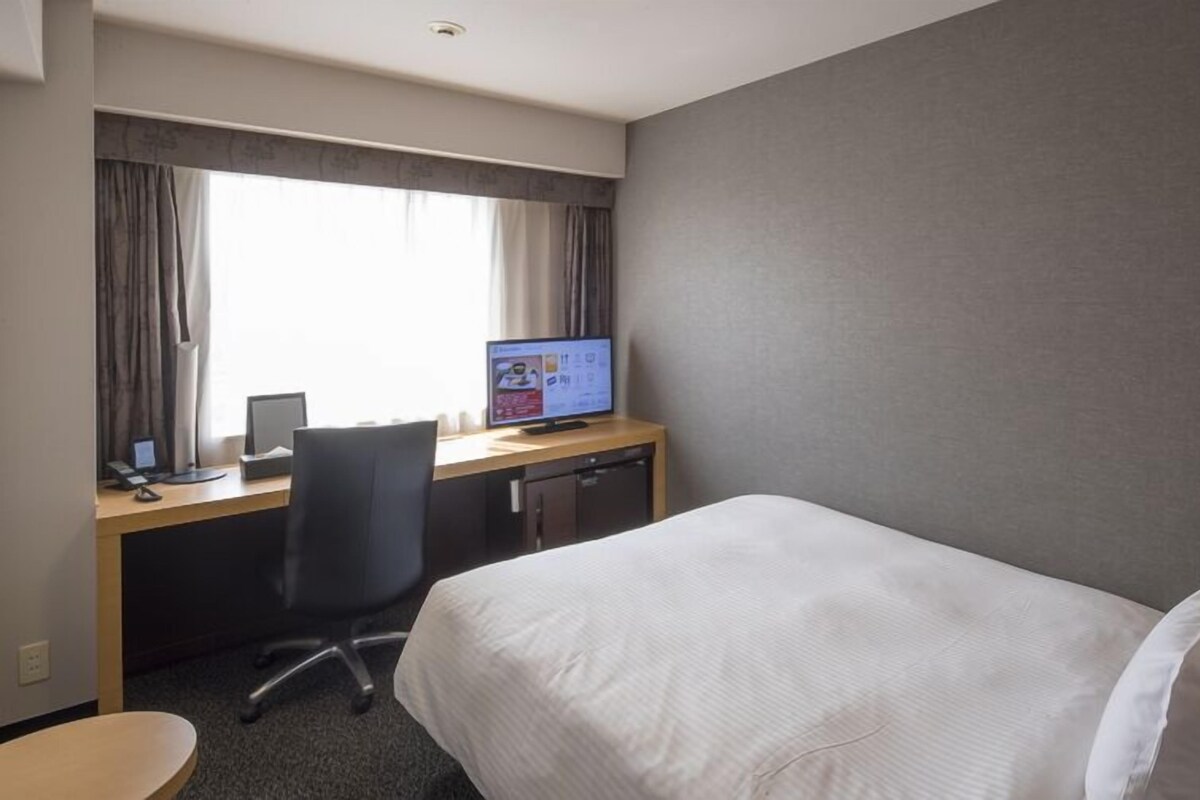 A hotel room with a bed, a desk with a computer, a chair, and a window with curtains.
