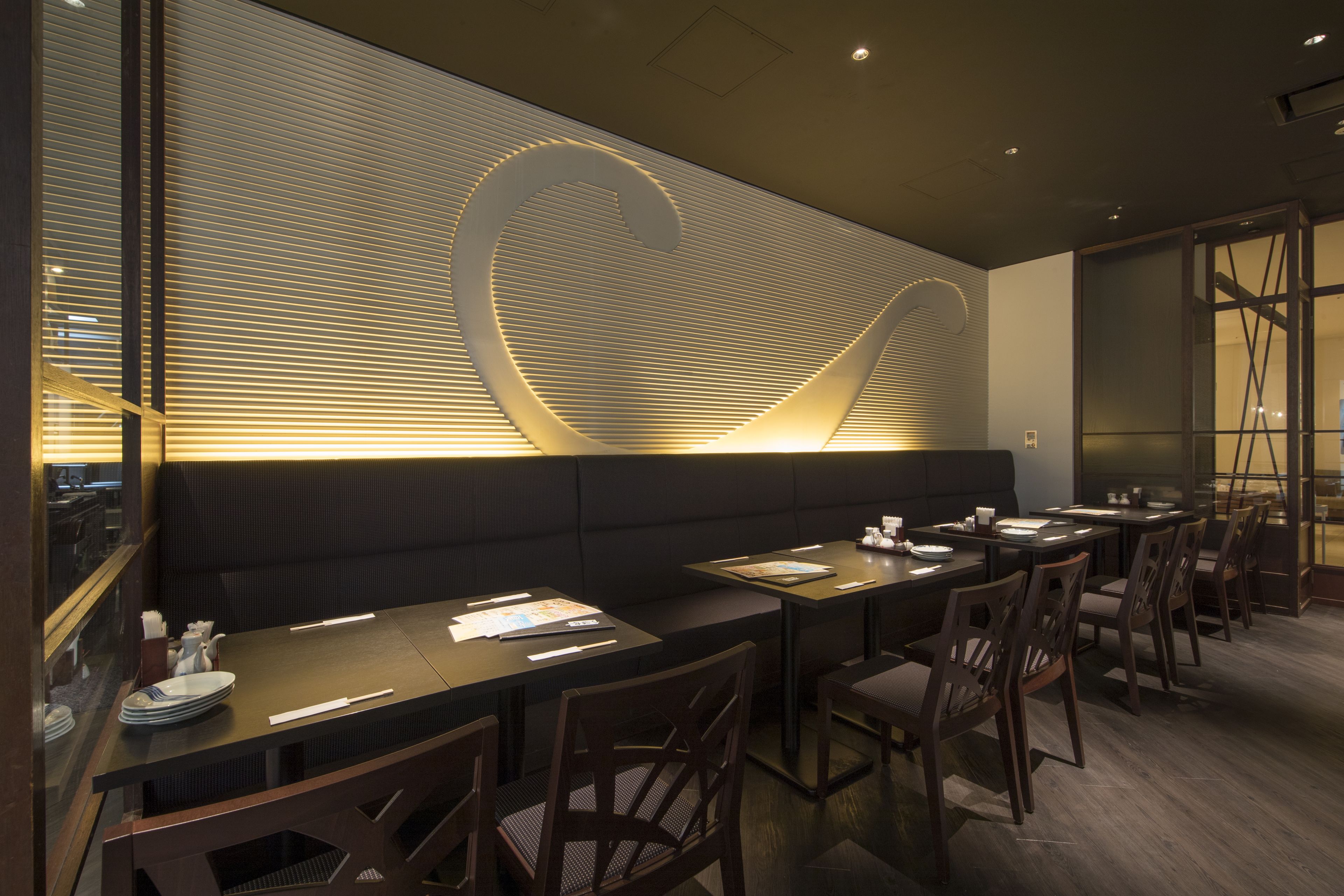 A modern restaurant interior with dark wood furniture, a large illuminated wall design, and a view of the city through large windows.