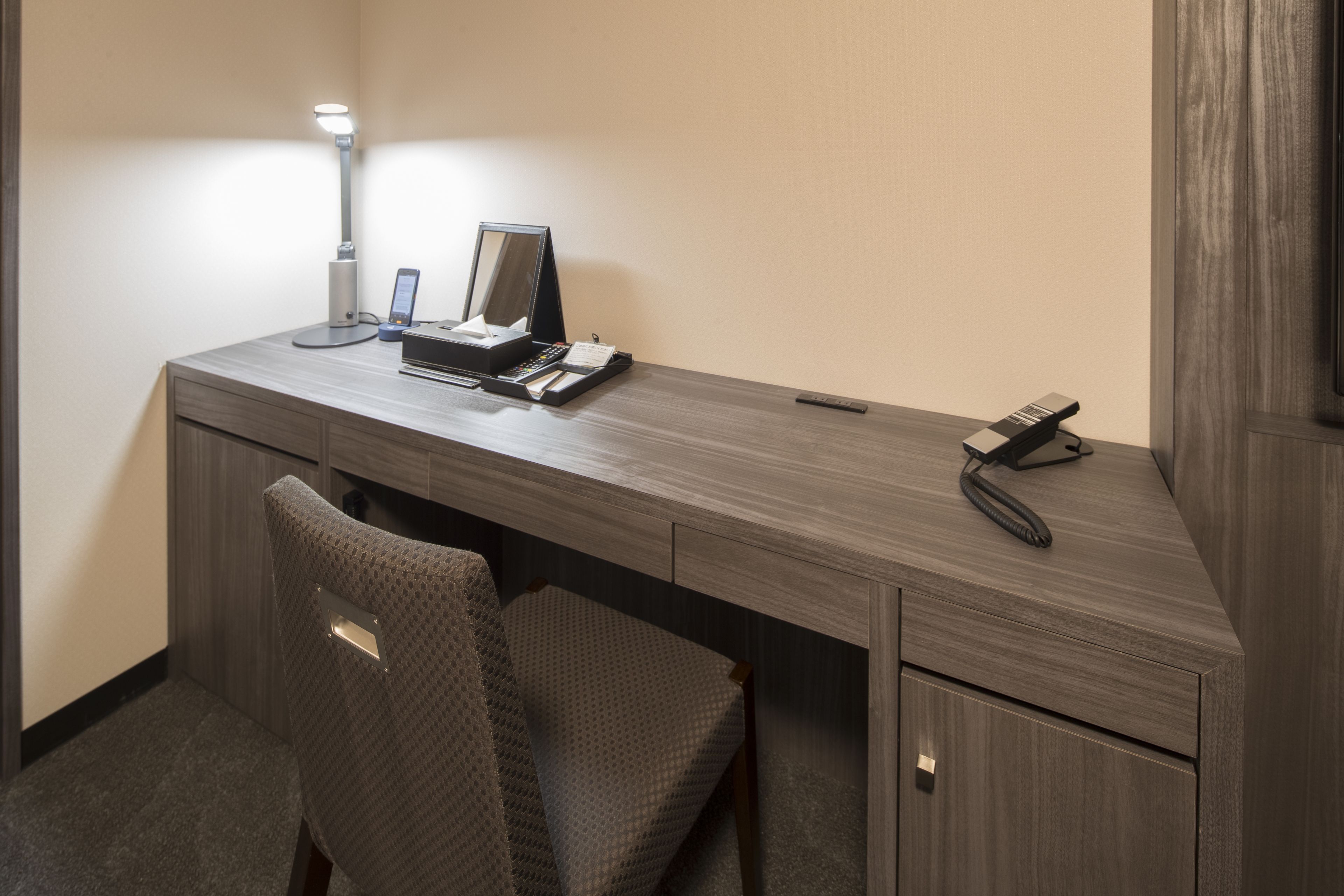 A hotel room with a wooden desk, a chair, a desk lamp, a phone, and a remote control.