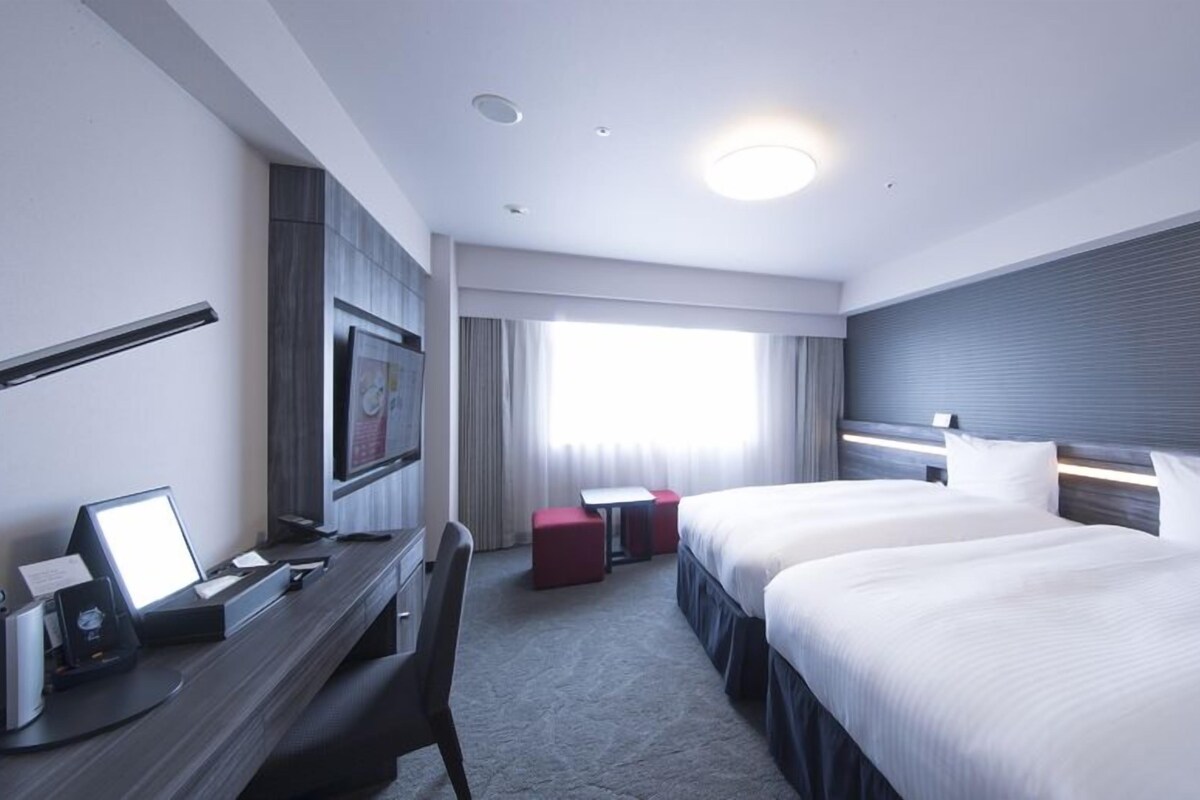 Habitación de hotel con dos camas, un escritorio con computadora, un televisor y una ventana con cortinas.