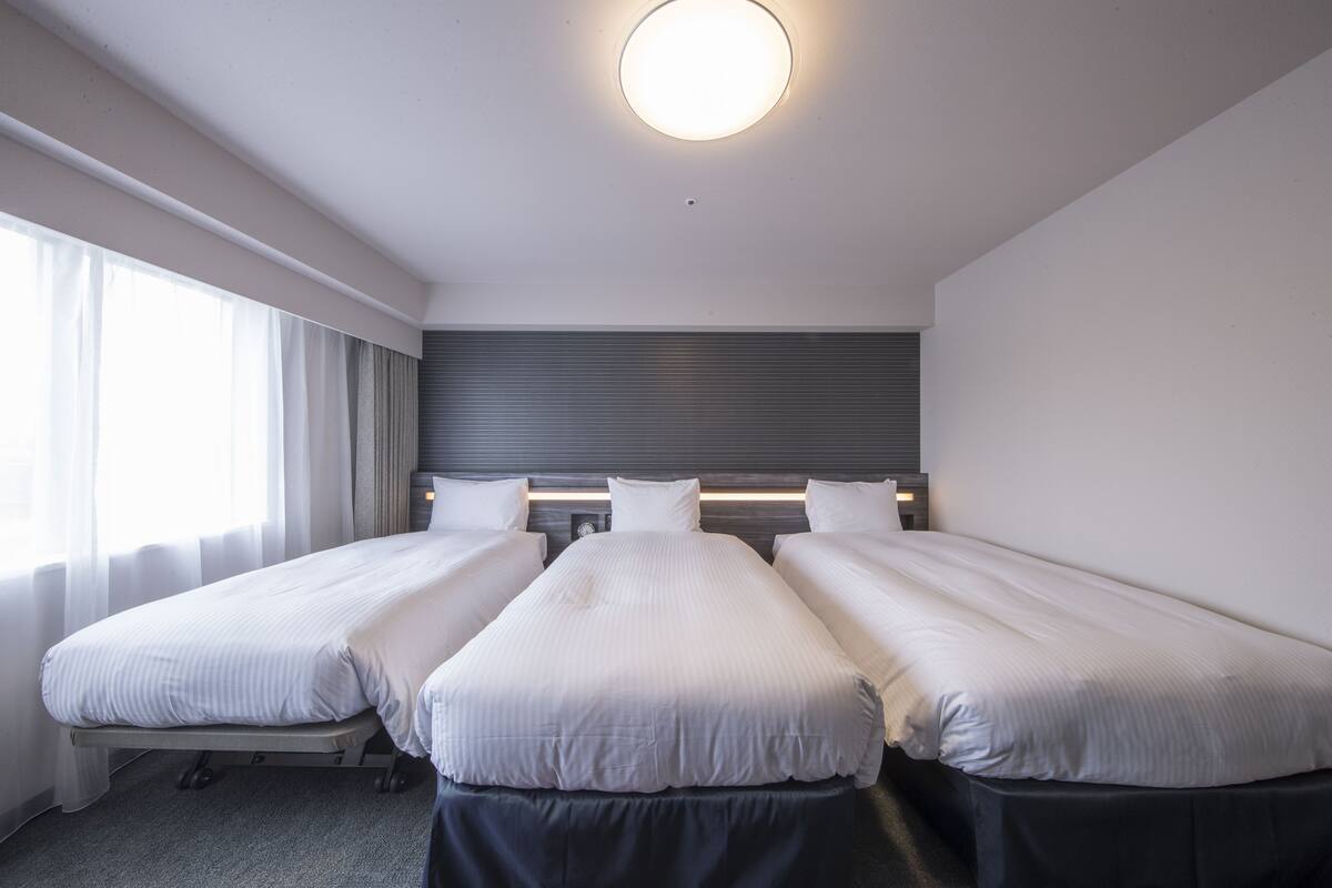 A hotel room with three single beds, a large window with curtains, and a modern ceiling light.
