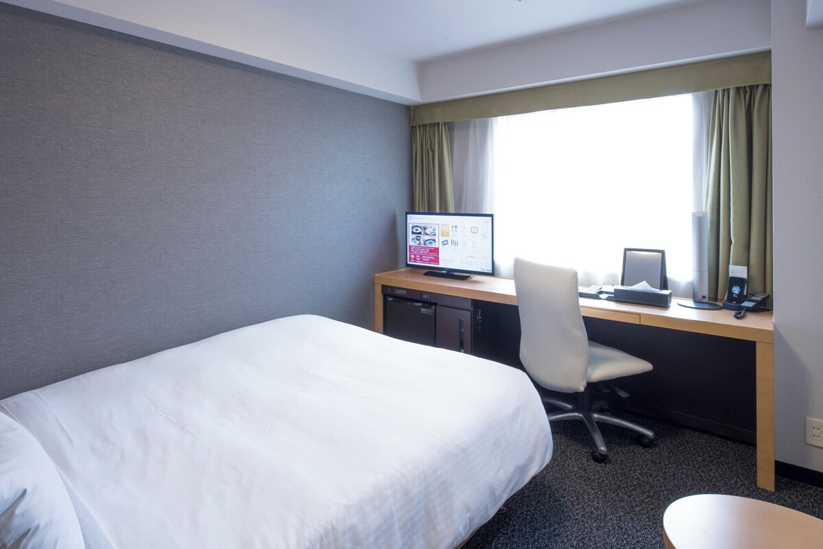 A hotel room with a bed, desk, computer, chair, and window with curtains.