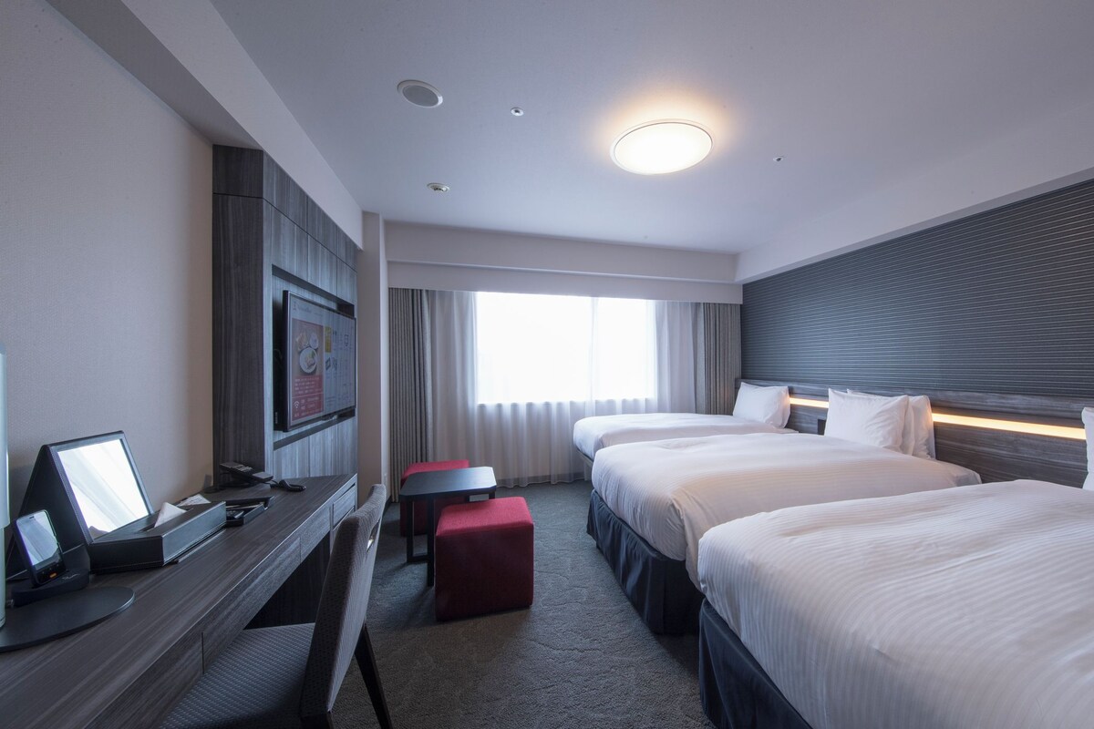 A hotel room with a desk, two beds, a TV, and a window with curtains.