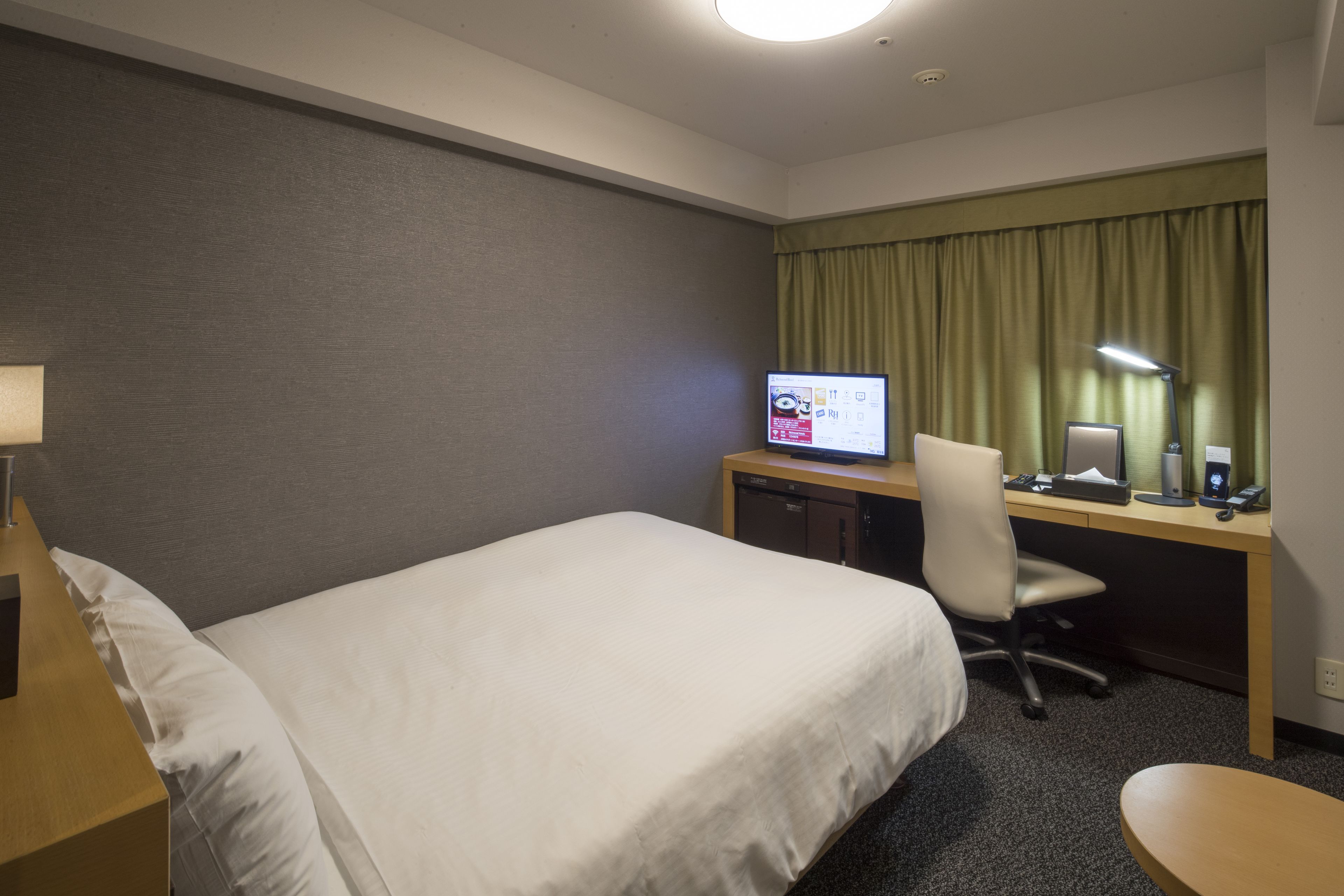 A hotel room with a single bed, a desk with a computer, a chair, and a lamp.