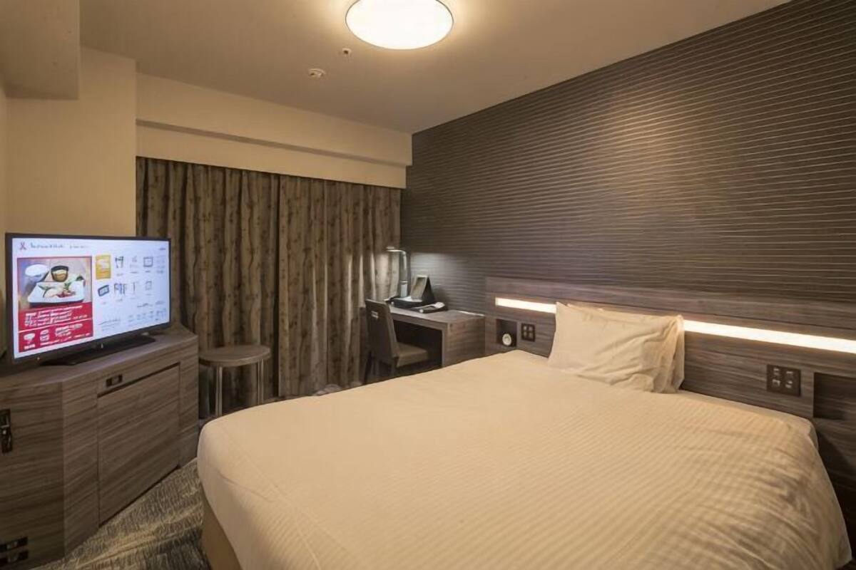 A hotel room with a bed, a desk with a computer, a TV, and a wall-mounted light.