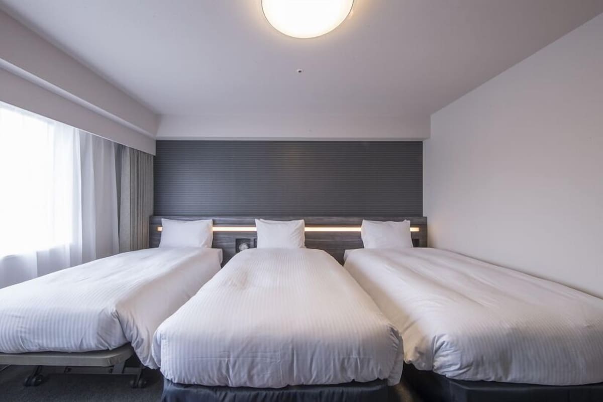 Habitación de hotel con tres camas individuales, un ventanal grande con cortinas, un cabecero moderno y una lámpara en el techo.