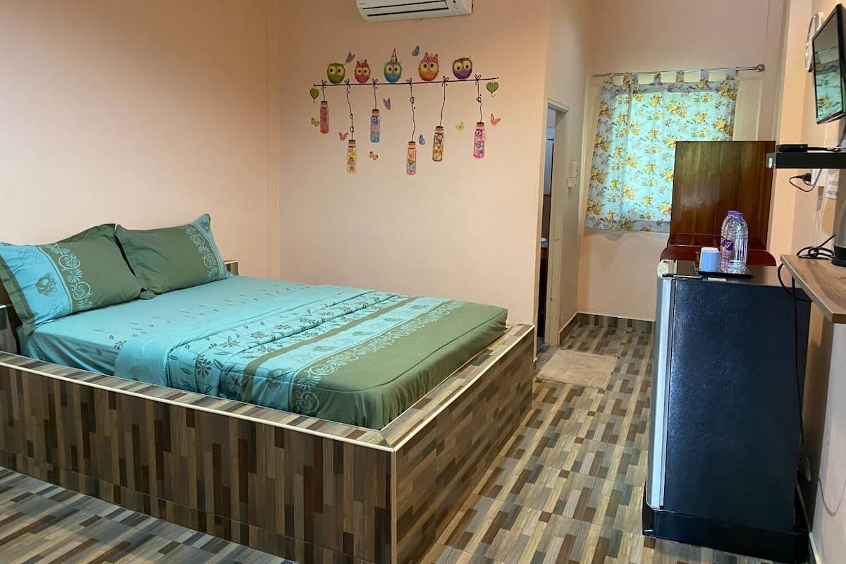 A bedroom with a bed, a wooden headboard, a wall-mounted air conditioner, a refrigerator, and a floral curtain.