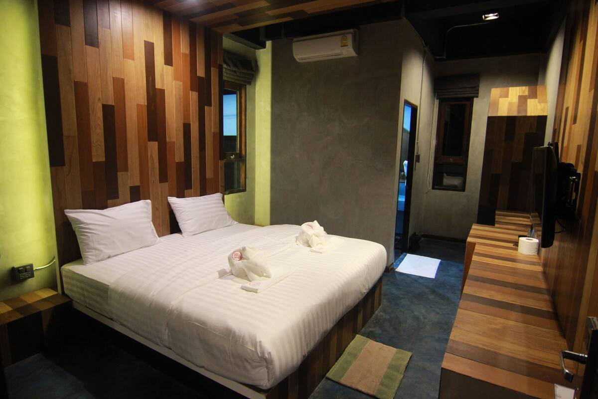 Habitación de hotel con cabecera de madera, una cama grande con ropa blanca, un banco de madera al pie de la cama y suelo de madera.