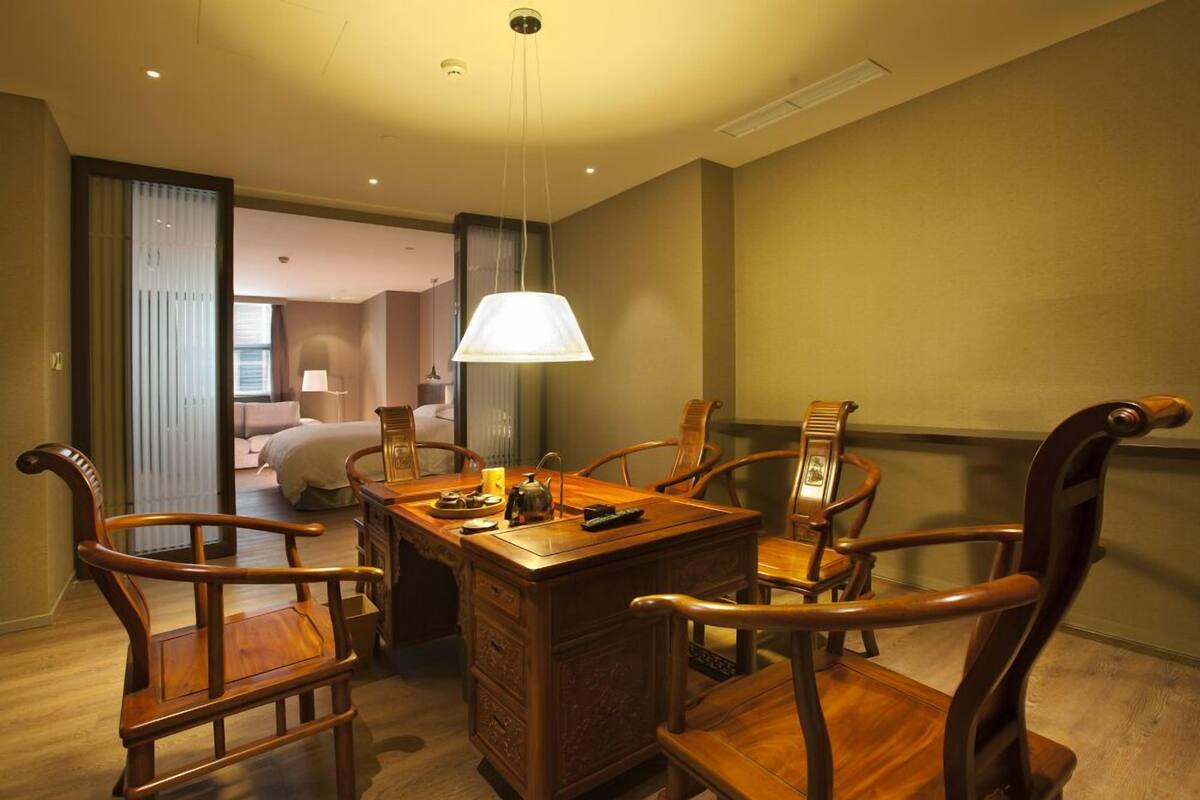 A hotel room with a wooden dining table, four chairs, a lamp, and a bed in the background.