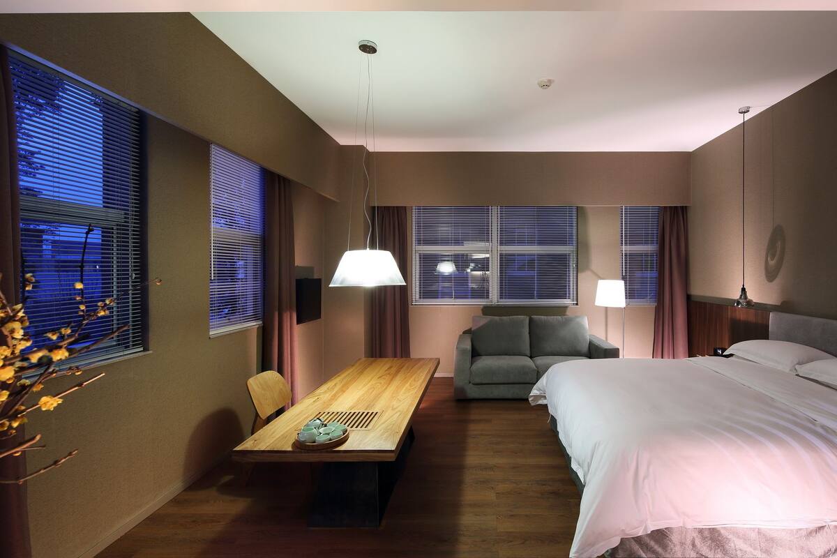 A hotel room with a bed, a sofa, a dining table, and two windows with blinds.