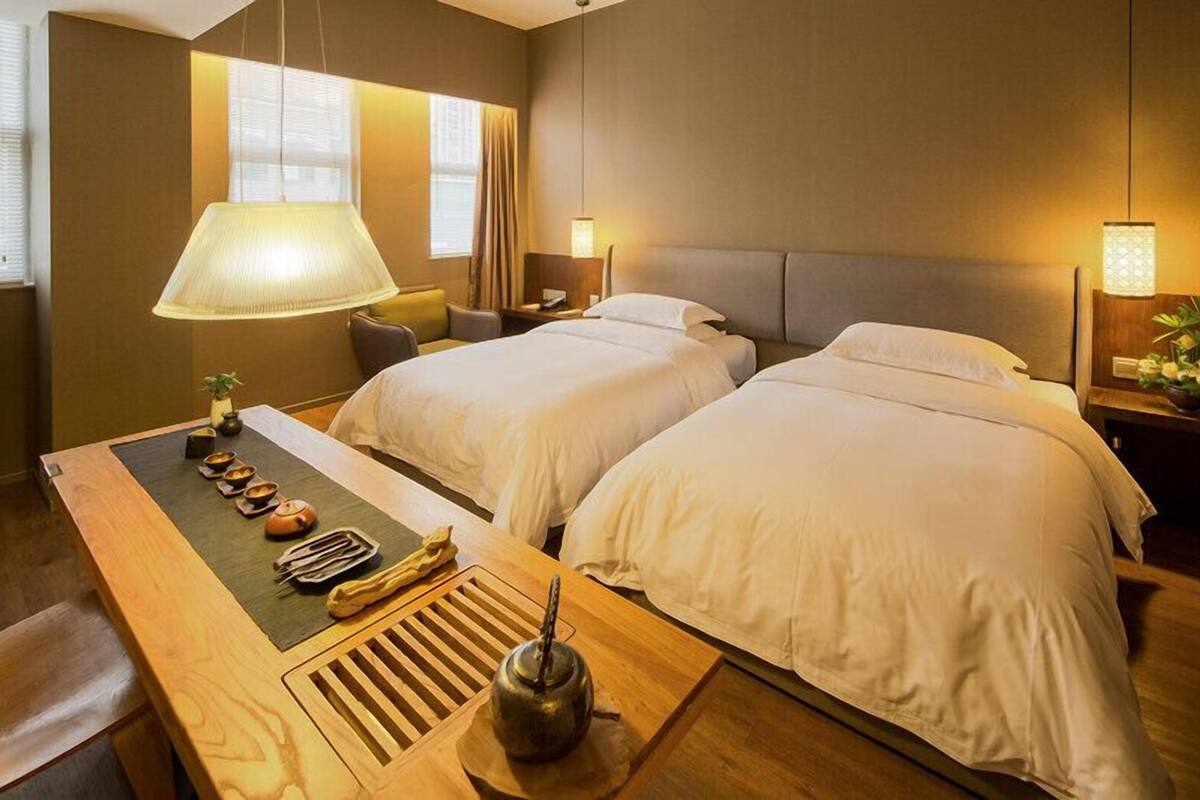 A hotel room with two beds, a wooden table with tea set, and a large window with curtains.